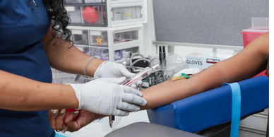 Medical Assistant draws blood from a patient's arm, who holds a red stress ball. Medical setting.